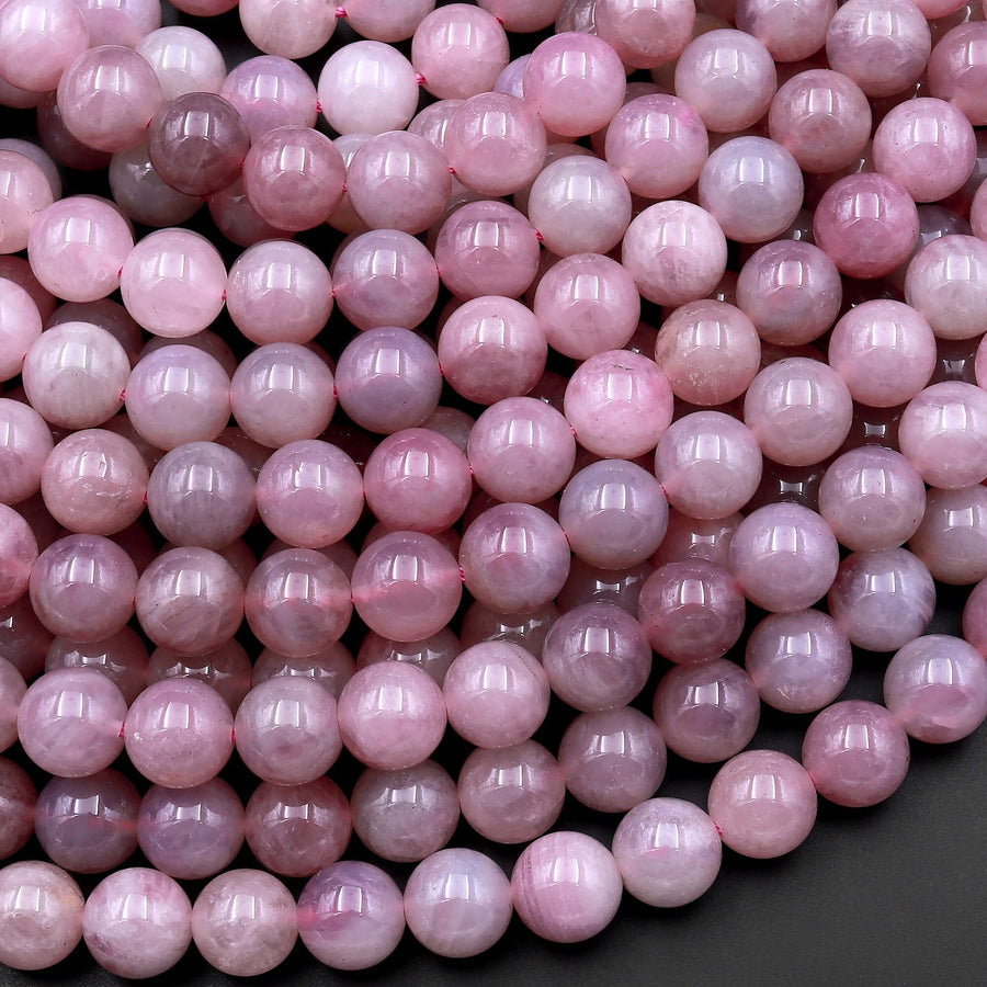 May include: A close-up image of a strand of pink gemstone beads. The beads are round and smooth, with a light pink color. The beads are arranged in a random pattern, and some of the beads have a slight shimmer to them.