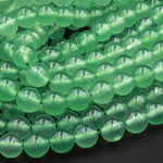 May include: A strand of smooth, round, green jade beads. The beads are translucent and have a slight shimmer. The beads are arranged in a loose pile, showing the individual beads.
