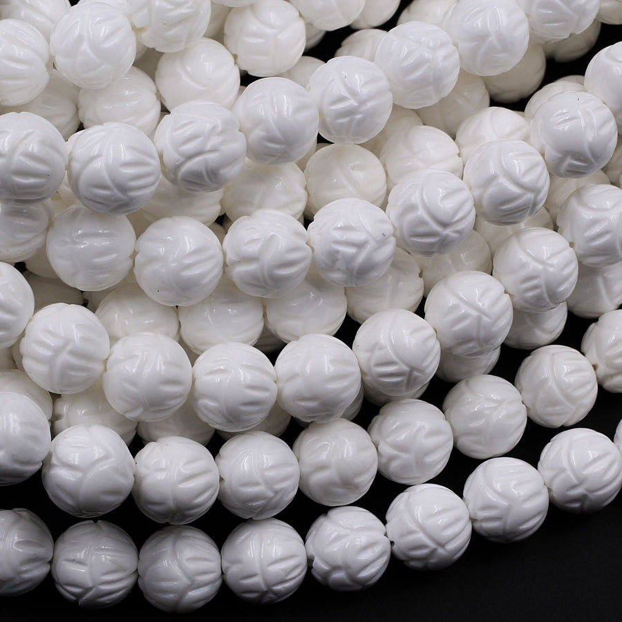 May include: A close-up image of a strand of white carved beads. The beads are round and have a floral design carved into them. The beads are arranged in a pile, showing the intricate detail of the carving.