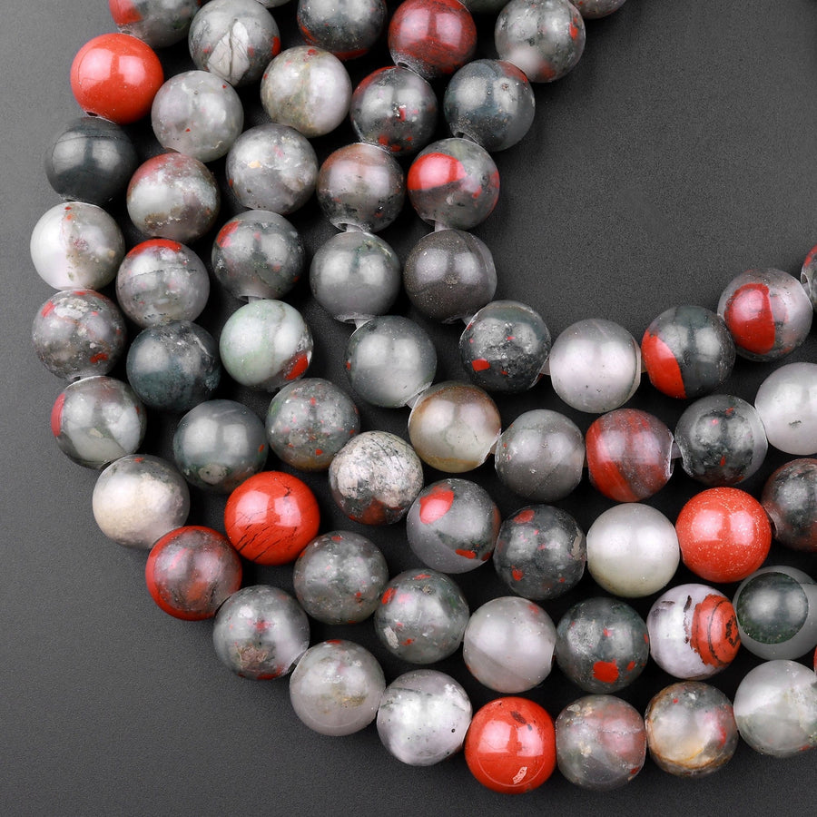 May include: A strand of round, gray and red gemstone beads. The beads are translucent and have a speckled pattern. The beads are arranged in a circular pattern, with the red beads interspersed throughout the gray beads.