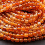 May include: A close-up image of a strand of orange carnelian beads. The beads are round and smooth, with a translucent, slightly cloudy appearance. The beads are arranged in a random pattern, creating a visually appealing texture.