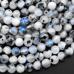 May include: A close-up of a pile of round, white moonstone beads with black and blue inclusions. The beads are smooth and polished, and they have a subtle iridescent sheen.