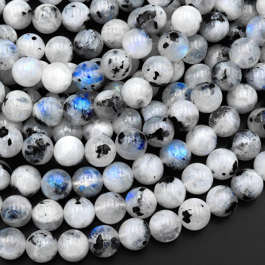 May include: A close-up of a pile of round, white moonstone beads with black and blue inclusions. The beads are smooth and polished, and they have a subtle iridescent sheen.