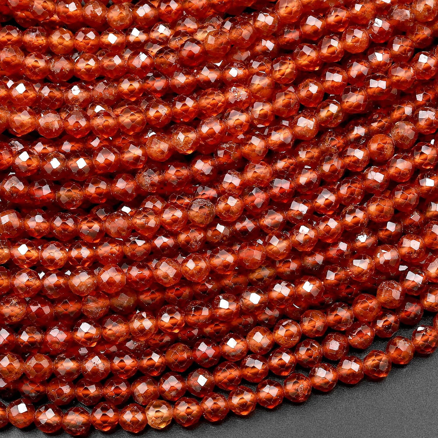 May include: A strand of faceted garnet beads. The beads are a deep reddish-orange color and have a glossy finish. The beads are arranged in a close-up view, showing the intricate facets of each bead.