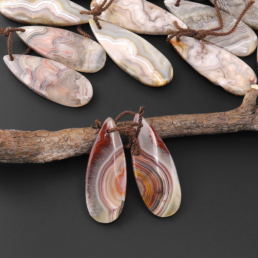May include: Two teardrop shaped agate pendants with brown string attached. The agate is a mix of white, brown, red, and orange colors with a swirling pattern.