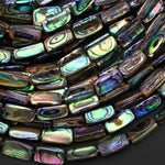 May include: A close-up of a pile of iridescent abalone shell beads. The beads are rectangular and have a variety of colors, including green, blue, purple, and brown. The beads are arranged in a random pattern and are reflecting light.