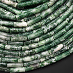 May include: A close-up of a strand of green and white jade beads. The beads are cylindrical and have a mottled, speckled appearance. The beads are arranged in a spiral pattern, creating a visual texture.