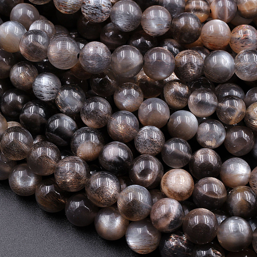 May include: A close-up image of a pile of round, black and silver sunstone beads. The beads are smooth and shiny, with a subtle shimmer. The beads are arranged in a random pattern, with some overlapping.
