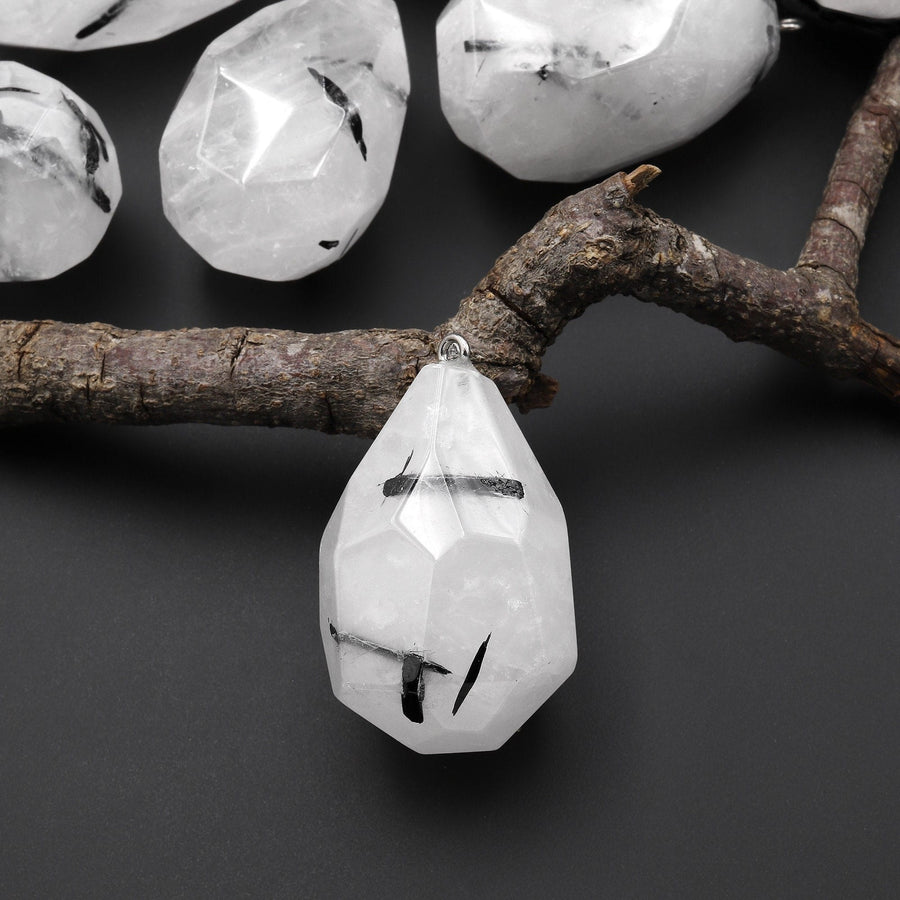 May include: A clear quartz pendant with black tourmaline inclusions, shaped like a teardrop, hanging from a silver chain. The pendant is resting on a branch.